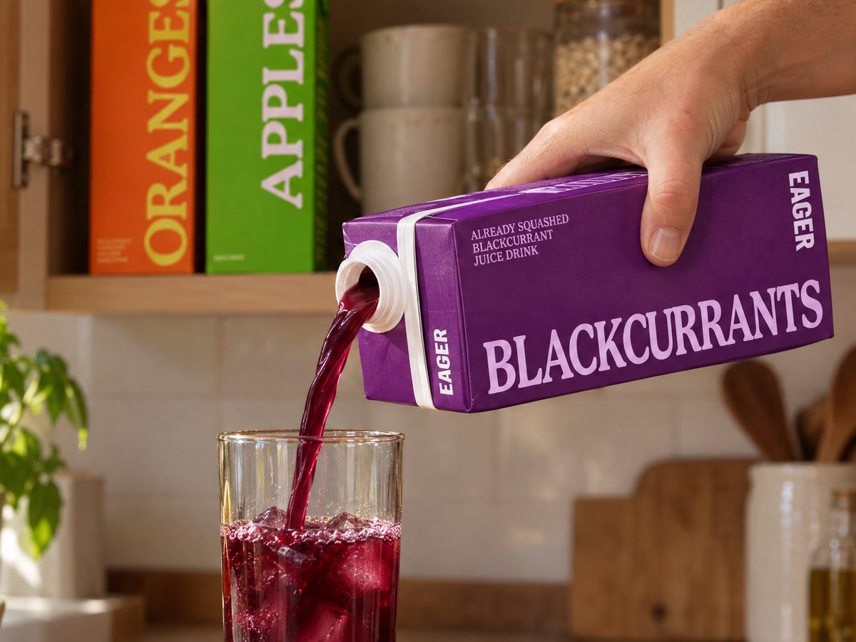 Dark purple juice poured from the Blackcurrants carton into a glass of ice, with Orange and Apple cartons on a cupboard shelf behind.